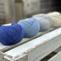 Four balls of wool in royal blue, medium denim blue, silvery grey, and cream. 