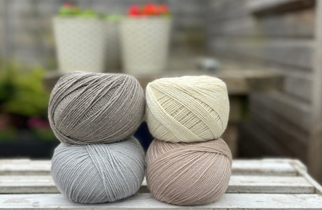 Four balls of wool in medium grey, cream, silvery grey, and oatmeal beige. 