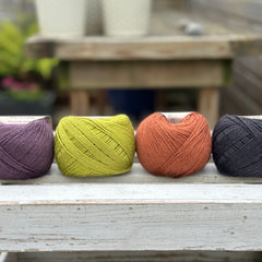 Four balls of wool in dark aubergine purple, earthy lime green, rust, and dark grey. 