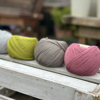 Four balls of wool in silvery grey, rich earthy lime green, medium grey, and medium soft pink. 