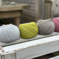 Four balls of wool in silvery grey, rich earthy lime green, medium grey, and medium soft pink. 