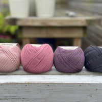 Four balls of wool in light warm pink, medium soft pink, dark aubergine purple, and dark grey. 