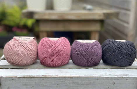 Four balls of wool in light warm pink, medium soft pink, dark aubergine purple, and dark grey. 