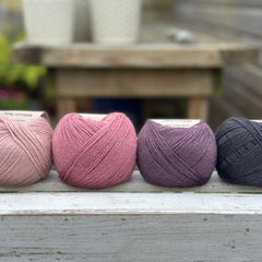 Four balls of wool in light warm pink, medium soft pink, dark aubergine purple, and dark grey. 