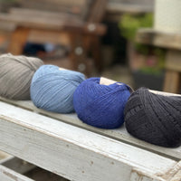 Four balls of wool in mid-grey, medium denim blue, royal blue, and dark grey. 