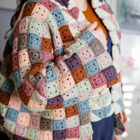 A person with long brown hair wearing a patchwork granny square jacket over blue dungarees